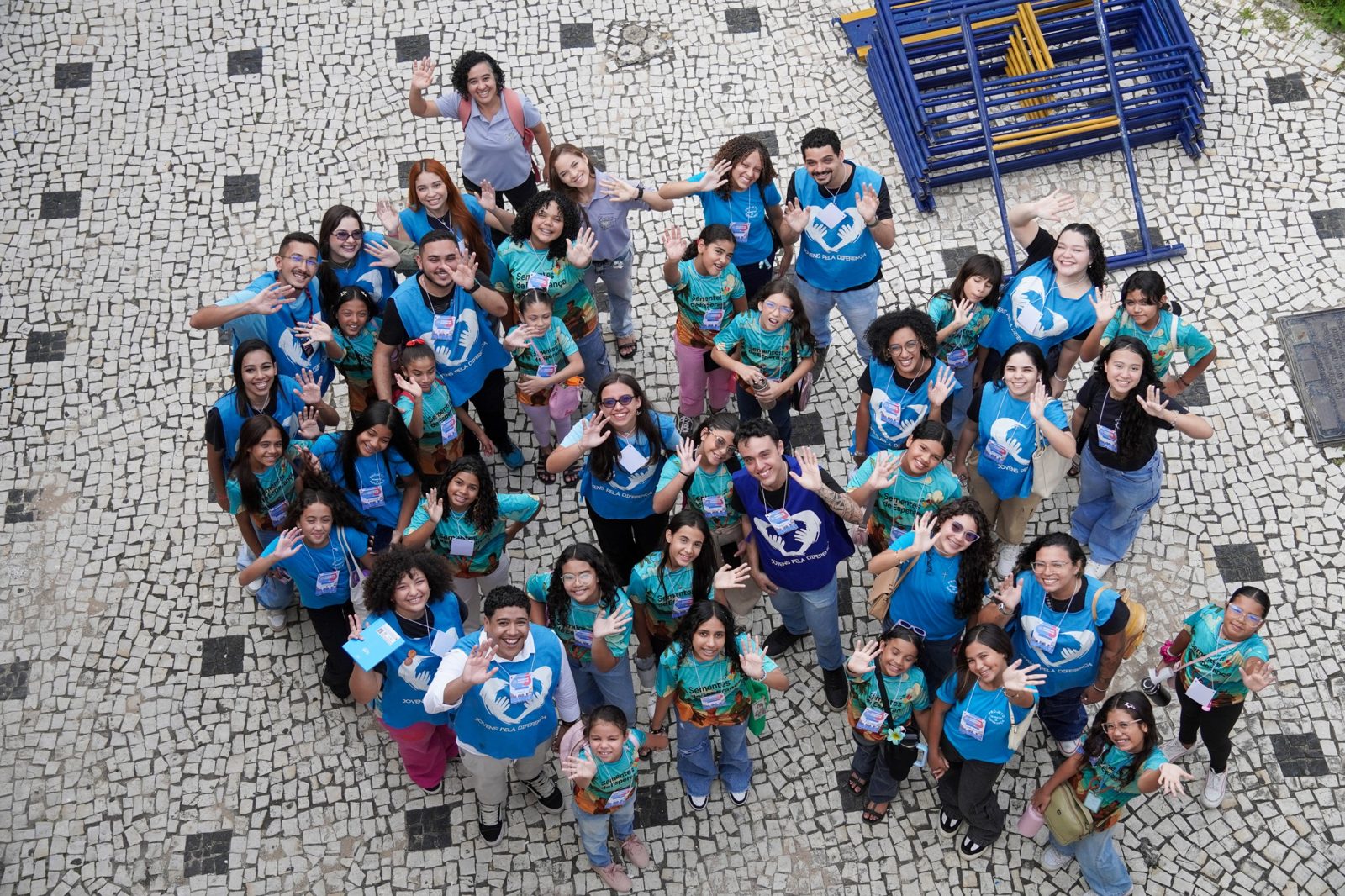 Voluntários do JPD com crianças, todos de colete e camiseta azul, acenando em um pátio de pedra portuguesa.