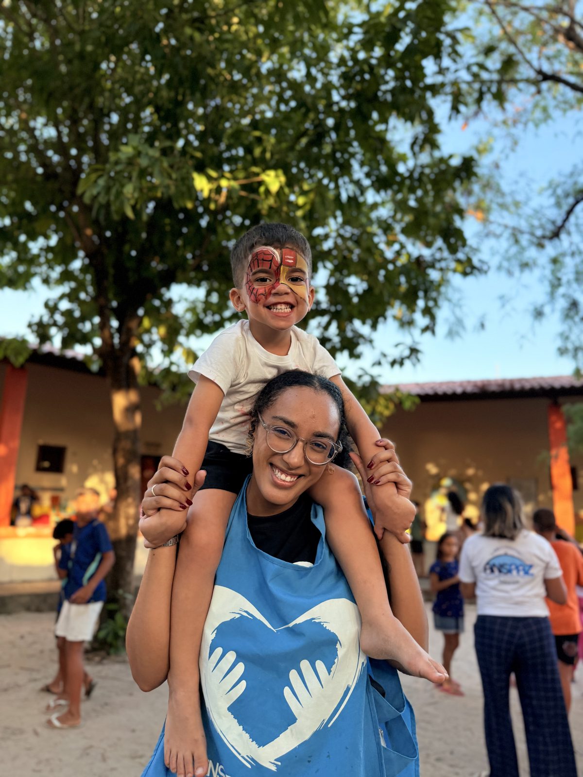 Voluntária sorrindo com uma criança de pintura facial nos ombros.