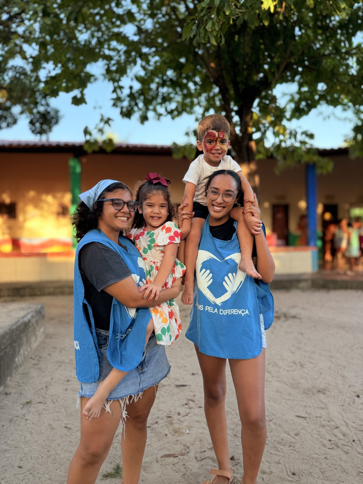 Duas voluntárias de colete azul com duas crianças em um pátio de escola.